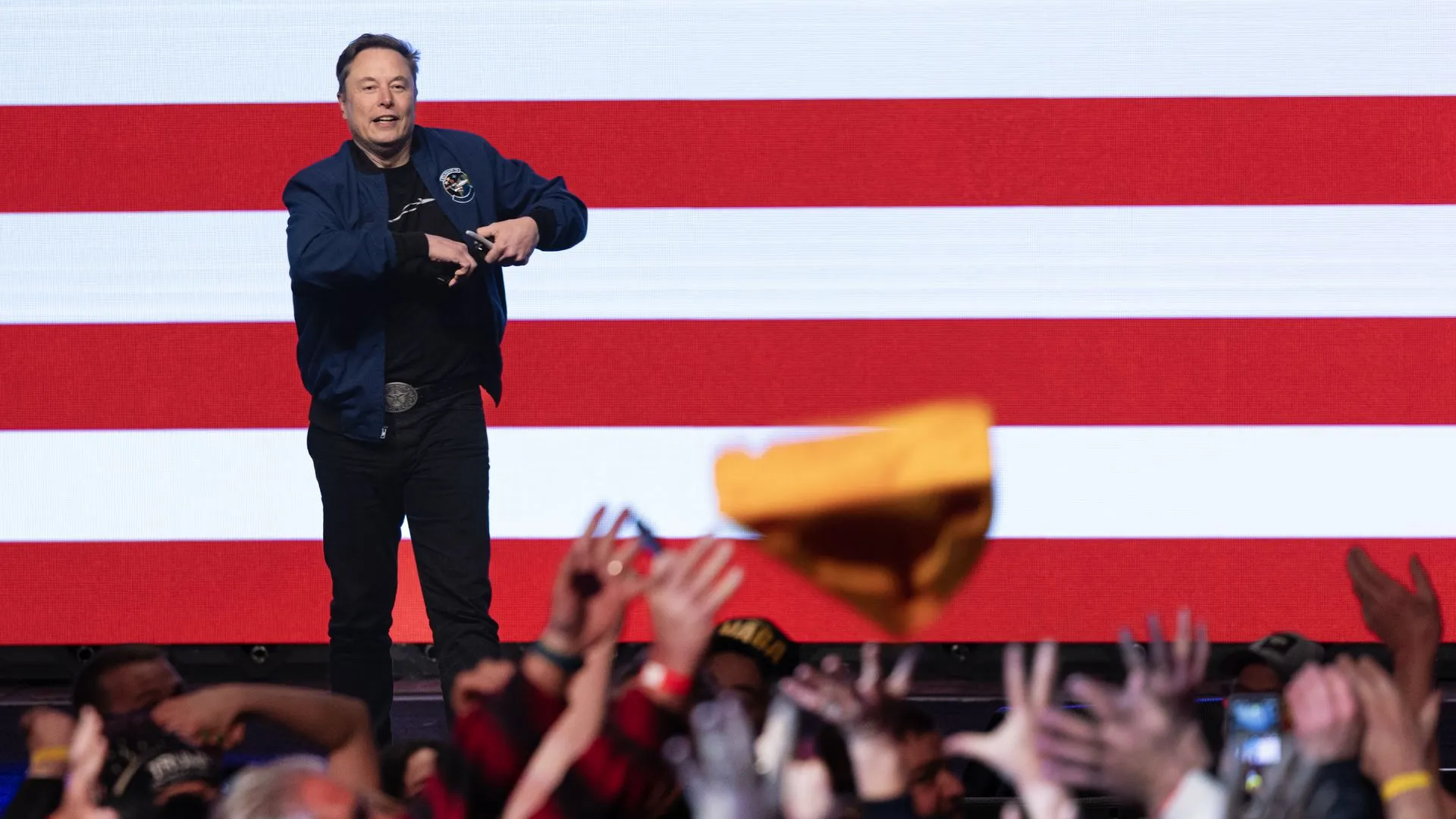 Elon Musk wearing a blue windbreaker and standing in front of an American flag wall.