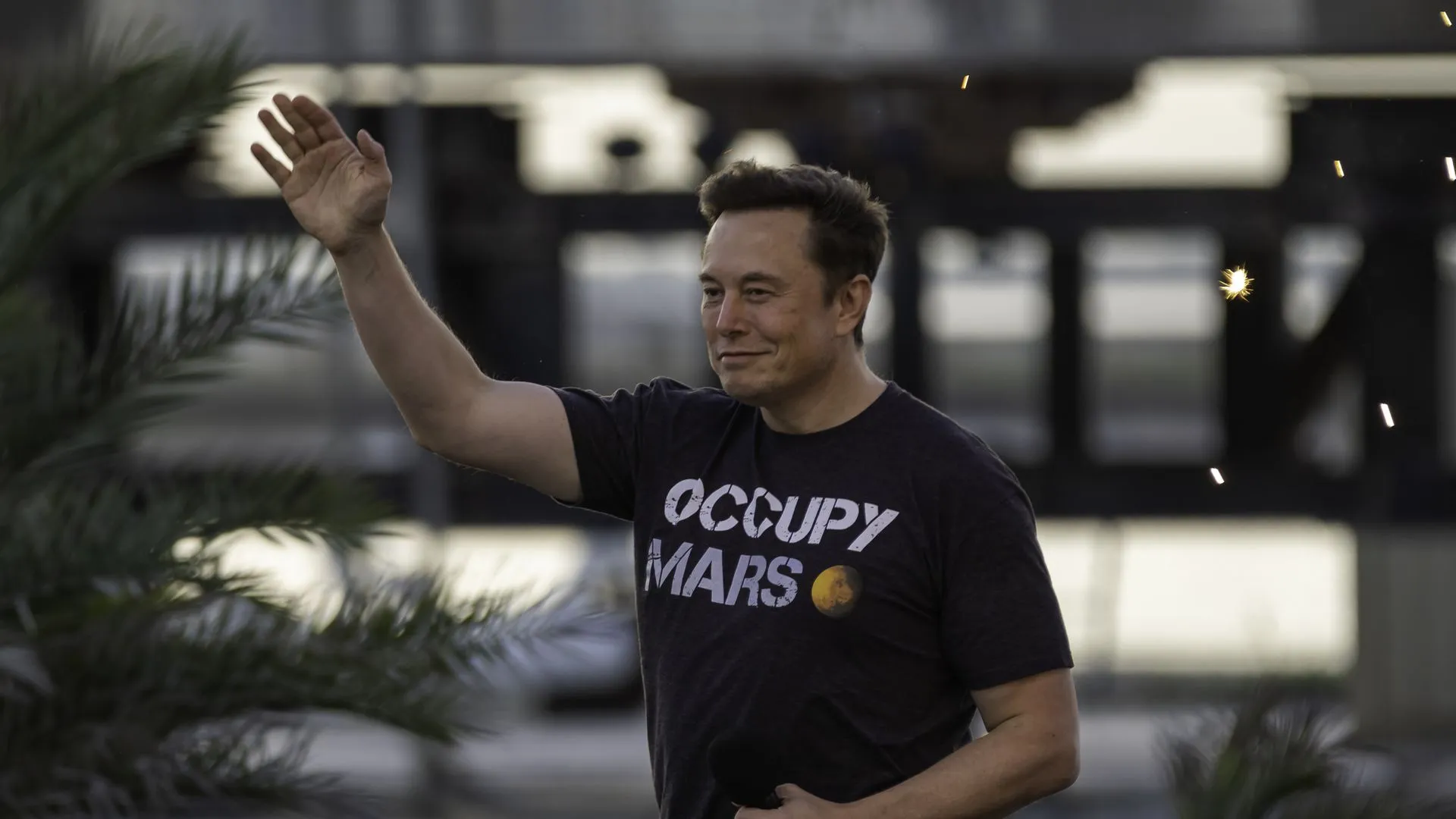 Elon Musk walks with a black shirt that reads "occupy Mars" and one arm outstretched 